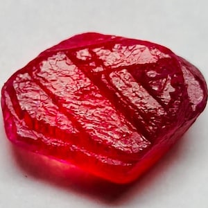 Könnte beinhalten: Ein facettierter, roher Rubin-Edelstein. Der Rubin hat eine tiefe, satte rote Farbe mit einer strukturierten Oberfläche. Der Edelstein ist unregelmäßig geformt und erscheint durchscheinend.