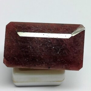 Puede incluir: Una gema rectangular y facetada de color burdeos intenso. La gema tiene una superficie pulida y reflectante con una sutil textura interna. Se muestra sobre una pequeña base rectangular blanca, resaltando su forma y color.