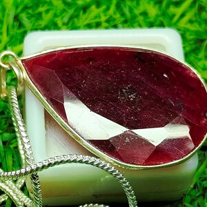 May include: A teardrop-shaped pendant with a faceted, deep red gemstone set in a gold-colored bezel. A silver-colored chain is attached to the pendant. The pendant is resting on a white surface, with a green background.