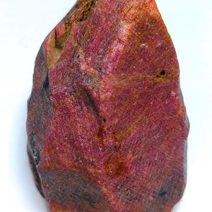 May include: A raw, uncut ruby gemstone, displaying a rich red colour with natural textures and variations. The ruby's surface shows a mix of dark and light red tones, with hints of orange and brown. The stone's irregular shape and raw appearance highlight its natural origin.