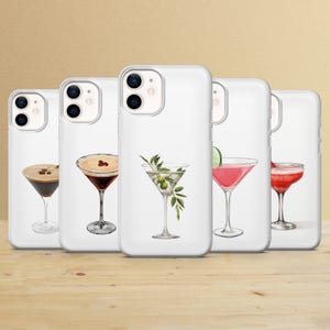May include: White phone cases with cocktail illustrations. The cases feature various drinks, including a martini with olives, a pink cocktail with a lime, and a coffee cocktail. The cases are arranged on a wooden surface.