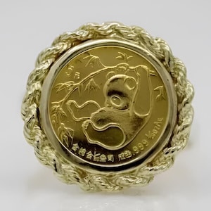 May include: A gold coin featuring a panda bear hanging from bamboo, set within a decorative, twisted gold border. The coin has Chinese characters and the text "999 Au". The coin is a collectible item.