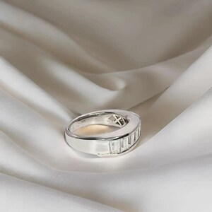 Pu&ograve; includere: Un anello d'argento con una fila di pietre preziose rettangolari e trasparenti. L'anello &egrave; posto su uno sfondo bianco e setoso. Il design dell'anello &egrave; moderno e minimalista, adatto per l'uso quotidiano o per occasioni speciali.