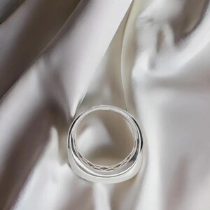Pu&ograve; includere: Un braccialetto argentato lucido dal design arrotondato. Il braccialetto &egrave; posto su uno sfondo di tessuto di raso bianco, creando un contrasto di trame e toni. La superficie liscia del braccialetto riflette la luce, evidenziando la sua finitura lucida.
