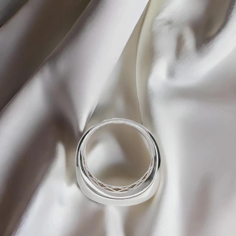 Pu&ograve; includere: Un braccialetto argentato lucido dal design arrotondato. Il braccialetto &egrave; posto su uno sfondo di tessuto di raso bianco, creando un contrasto di trame e toni. La superficie liscia del braccialetto riflette la luce, evidenziando la sua finitura lucida.