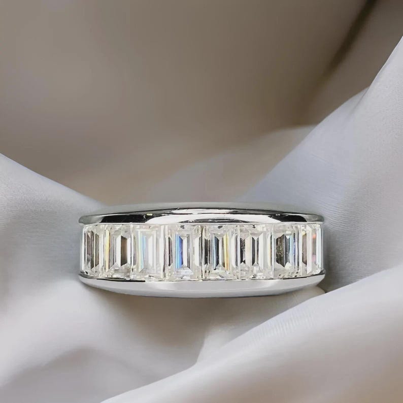 Pu&ograve; includere: Un anello d'argento con una fascia di pietre preziose rettangolari e trasparenti. L'anello &egrave; posto su un tessuto bianco drappeggiato, creando un contrasto che ne esalta la lucentezza e il design. Le pietre sono uniformemente distanziate.