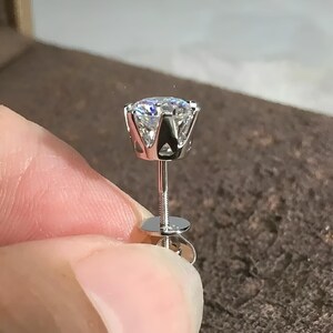 May include: A close-up of a single, sparkling diamond stud earring. The diamond is round-cut and set in a silver prong setting. The earring post and backing are also silver. The earring is held up by a person's fingers.