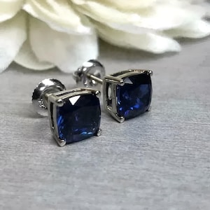 May include: A pair of square-cut, dark blue gemstone stud earrings with a silver setting. The earrings are displayed on a gray surface, with a blurred white flower in the background. The gemstones have a slight sparkle.