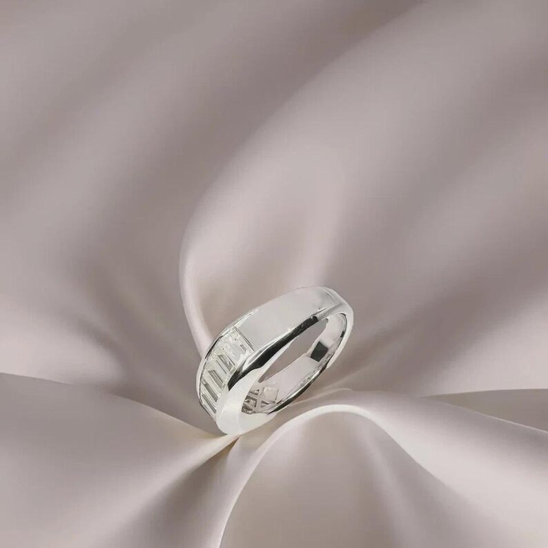 Pu&ograve; includere: Un anello d'argento con una fascia rettangolare e una fila di pietre a taglio rettangolare. L'anello &egrave; posto su uno sfondo di tessuto morbido e fluido.