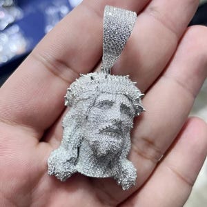 以下が含まれることがあります： 小さな輝く石で覆われた、イエスの顔を描いたシルバーペンダント。ペンダントは、いばらの冠を含む詳細なデザインです。ベイルも石で覆われています。ペンダントは人の手で持たれています。