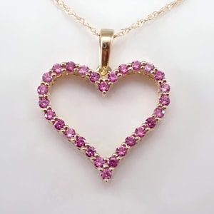 Könnte beinhalten: Eine goldfarbene Herz-Anhänger-Halskette. Das Herz ist mit kleinen, runden, rosa Edelsteinen umrandet. Die Halskette hat eine zarte, gedrehte Kette.