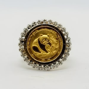 May include: A gold ring featuring a gold coin with a panda bear design. The coin is surrounded by a halo of small, clear gemstones set in a silver-colored metal. The ring is set against a white background.