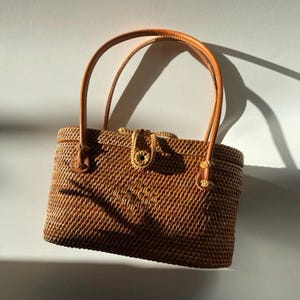May include: A rectangular woven rattan handbag with a warm brown finish. The bag features two smooth, rounded leather handles attached to the top, and a central woven closure tab with a small circular button detail at the front center.