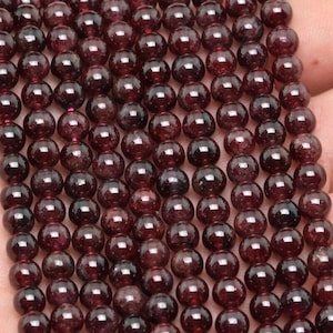 Könnte beinhalten: Nahaufnahme von Strängen polierter, runder, dunkelroter Granatperlen. Die Perlen sind aufgefädelt, ideal für die Schmuckherstellung. Die Perlen haben eine glänzende Oberfläche und sind gleichmäßig groß.