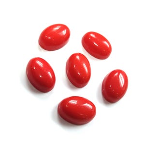 May include: Six oval-shaped, polished red gemstones are arranged on a white background. Each stone has a smooth, glossy surface, reflecting light. The gemstones are a vibrant, solid red color, and appear to be of similar size and shape.