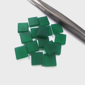 May include: A collection of small, square, translucent green gemstone cabochons arranged in a cluster. A pair of silver-toned metal tweezers is positioned diagonally across the top right corner, providing a sense of scale for the stones.