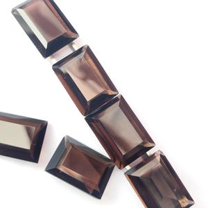 May include: A collection of five rectangular, faceted smoky quartz gemstones. Each stone features a deep brown, translucent color with sharp, clean edges and a polished surface, arranged in a linear pattern with two stones placed separately.