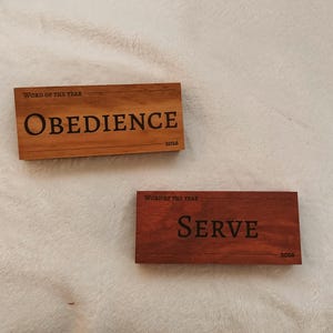 May include: Two rectangular wooden plaques. The top plaque is light brown and reads "WORD OF THE YEAR" above the word "OBEDIENCE" in large, dark letters. The bottom plaque is dark red and reads "WORD OF THE YEAR" above the word "SERVE". Both have "2026" at the bottom.