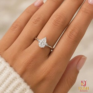 May include: A silver-toned ring featuring a central pear-cut clear gemstone flanked by two smaller round-cut stones on a slender band. The jewelry piece is marked with the logo EJ Elish Jewels in the bottom right corner of the image.