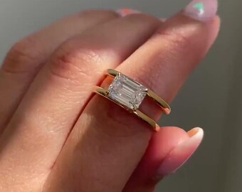 May include: A gold ring featuring a rectangular, clear gemstone. The ring's band has a unique open design, with the gemstone held between two parallel bars. The ring is shown on a hand with light pink nail polish.
