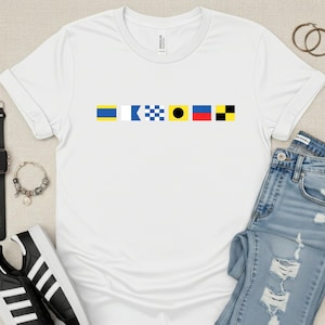 May include: White t-shirt with a nautical flag design in blue, yellow, red, and black. The shirt is paired with light blue denim jeans, black and white sneakers, a black watch, and gold hoop earrings. A silver bracelet with charms is also visible.