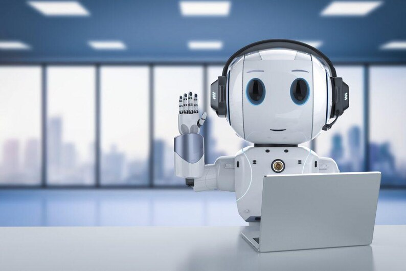 Op de afbeelding: Een witte robot met een headset en een laptop, die zijn rechterhand opheft als begroeting. De robot heeft grote, donkere ogen en een vriendelijke uitdrukking. De achtergrond is een vage kantooromgeving met grote ramen.