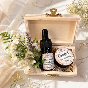 Puede incluir: Una caja de madera clara con un cierre dorado contiene una botella negra de aceite corporal "Sensuality" y un tarro de manteca corporal "Comfort". La caja está decorada con flores blancas y moradas.