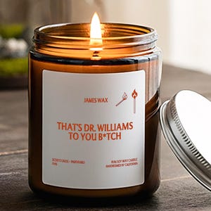 May include: A lit candle in an amber glass jar with a silver metal lid. The white label features the text 'JAMES WAX', an illustration of two matches, and the text 'THAT'S DR. WILLIAMS TO YOU B*TCH', along with smaller text at the bottom.