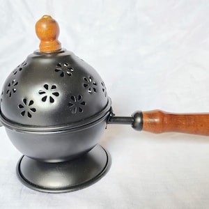 以下が含まれることがあります： 木製のハンドルと上部が付いた黒い金属製のお香バーナー。 球状の本体には花型の切り抜きがあります。 ハンドルと上部は明るい茶色の木材で作られています。 バーナーは丸いベースにあります。