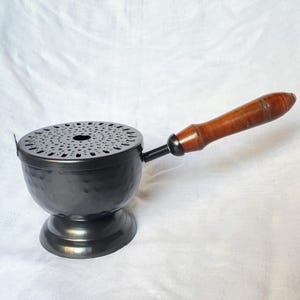 以下が含まれることがあります： 木製のハンドルが付いた、濃い灰色の金属製のお香立て。バーナーには、丸い穴あき蓋と、ボウルにハンマーで叩いたような質感があります。ハンドルは明るい茶色です。