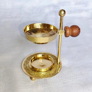 Puede incluir: Un quemador de incienso de latón con base redonda y cuenco perforado. La base tiene un diseño floral en relieve. Un pomo de madera está unido al lateral. El quemador está diseñado para contener incienso.
