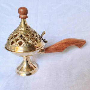 Może przedstawiać: Mosiężny palnik do kadzideł z drewnianym uchwytem i ozdobną pokrywą w romby. Palnik ma okrągłą podstawę i stożkową górę, z małym drewnianym guzikiem na górze. Uchwyt jest zakrzywiony i wykonany z jasnobrązowego drewna.