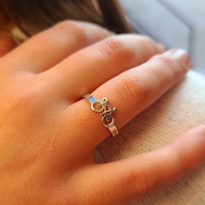 May include: A silver ring with a bicycle motif. The ring has a plain band with a small bicycle outline as the main feature. The bicycle has two wheels, a frame, and handlebars. The ring is worn on a finger.