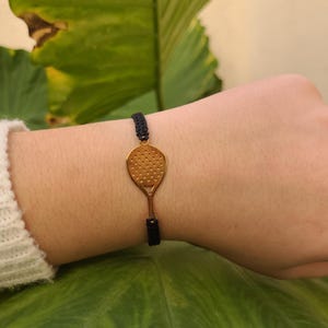 Puede incluir: Pulsera con un dije de raqueta de tenis dorada y un cordón negro tejido. El dije de la raqueta tiene una cara perforada y un mango delgado. La pulsera se lleva en una muñeca, con una manga de suéter blanca visible.