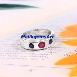 May include: A silver-toned ring featuring three circular gemstones set horizontally along the band. The centre stone is a vibrant red, flanked by two smaller deep blue stones. The text MalikgemsArt is printed in blue across the centre of the ring.