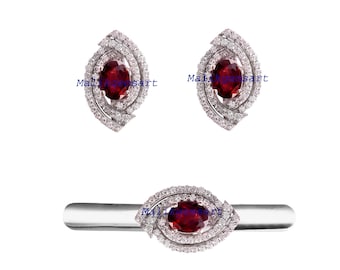 May include: A jewelry set featuring a pair of earrings and a ring. Each piece has a central oval, deep red gemstone surrounded by a halo of small, clear stones. The earrings and ring are set in a silver-colored metal.