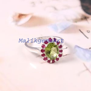May include: A silver ring featuring a central oval-cut light green peridot gemstone surrounded by a halo of small, round, deep red ruby stones. The text MalikgemsArt is overlaid in blue across the center of the ring band and the gemstone.