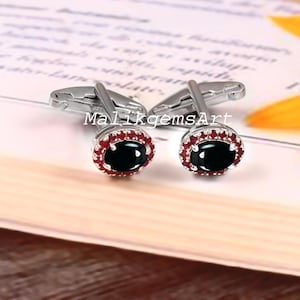 May include: Silver cufflinks featuring an oval, black gemstone encircled by small, red gemstones. The cufflinks are presented on a book, with the text "MalikgemsArt" visible.