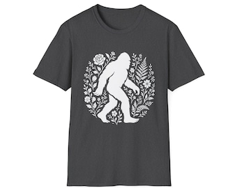 Puede incluir: Camiseta gris oscuro con una silueta blanca de Bigfoot dentro de una corona floral. El diseño incluye una rosa, helechos y otros elementos botánicos. La camiseta tiene cuello redondo y mangas cortas.