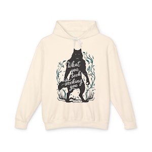 May include: A cream-coloured hoodie with a black wolf-like creature silhouette and the text "What you seek is seeking you." The design is framed by leaves. The hoodie has a drawstring hood.