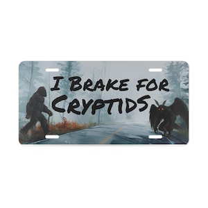 Puede incluir: Placa de matrícula rectangular con una escena de carretera y un bosque brumoso. El texto "I Brake for Cryptids" está en negro. Bigfoot está a la izquierda y una criatura parecida a Mothman está a la derecha.