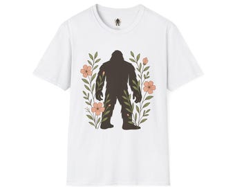 Puede incluir: Camiseta blanca con una silueta marrón oscuro de Bigfoot, flores color melocotón y hojas verdes. La camiseta tiene cuello redondo y mangas cortas. Un pequeño gráfico de Bigfoot está en la parte superior de la espalda.