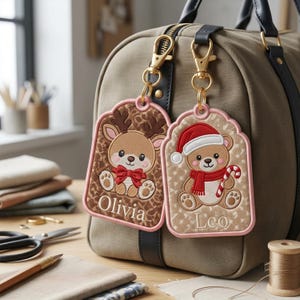Pode incluir: Duas etiquetas de bagagem bordadas com clipes de metal dourado presas a uma bolsa de lona. Uma etiqueta apresenta uma rena com um laço vermelho e o nome Olivia. A outra mostra um ursinho com gorro de Natal e o nome Leo.