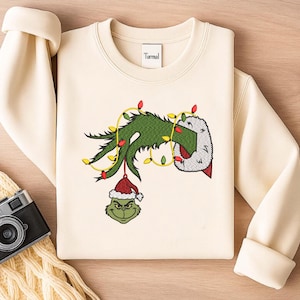 May include: Cream-colored sweatshirt featuring an embroidered Grinch design. The design includes the Grinch's green hand holding Christmas lights and a Grinch head wearing a Santa hat. The brand name "Turmal" is embroidered on the neckline.