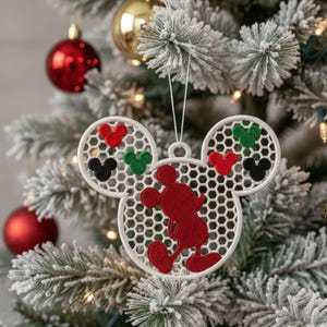 Könnte beinhalten: Ein weißer, gestickter Weihnachtsschmuck in Form von Mickey Mouse. Der Schmuck zeigt eine rote Mickey Mouse Silhouette und drei kleinere Kreise mit roten, grünen und schwarzen Mickey Mouse Kopfformen. Er hängt an einem verschneiten Weihnachtsbaum.