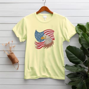 May include: A light yellow t-shirt featuring a graphic of an American flag and a bald eagle. The flag is red, white, and blue, and the eagle has brown and white feathers with a yellow beak. The shirt is hanging on a wooden hanger.