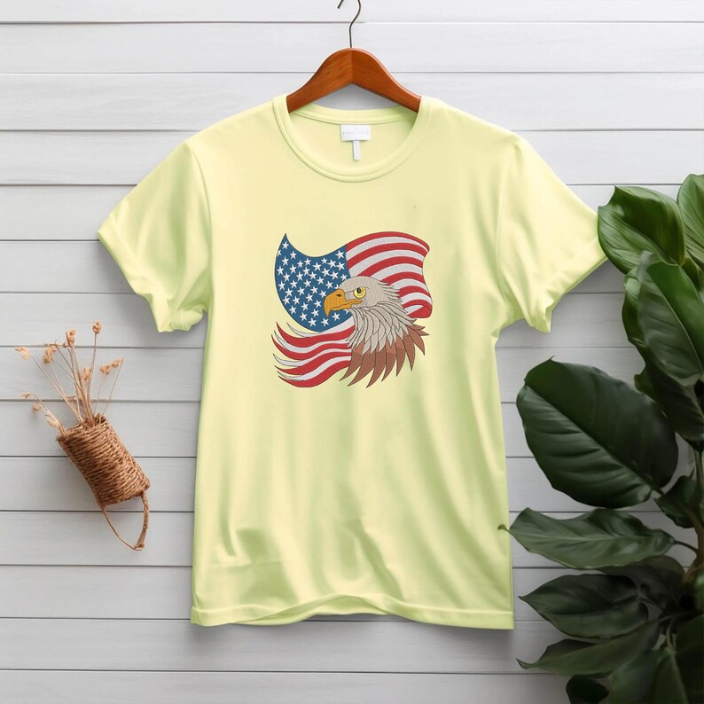 May include: A light yellow t-shirt featuring a graphic of an American flag and a bald eagle. The flag is red, white, and blue, and the eagle has brown and white feathers with a yellow beak. The shirt is hanging on a wooden hanger.