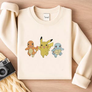 Może przedstawiać: Kremowa bluza z nadrukiem trzech postaci z kreskówek: Charmandera, Pikachu i Squirtle. Postacie są w kolorze pomarańczowym, żółtym i niebieskim. Nazwa marki "Turmal" jest wydrukowana na kołnierzu.