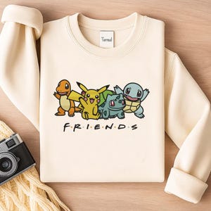 Könnte beinhalten: Cremefarbenes Sweatshirt mit einer Grafik von vier Zeichentrickfiguren und dem Wort "FRIENDS" in Schwarz. Die Figuren sind orange, gelb, grün und blau. Das Sweatshirt hat einen Rundhalsausschnitt und lange Ärmel.