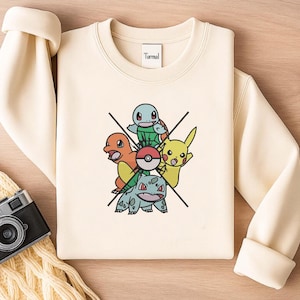 May include: Cream-coloured sweatshirt with a colourful graphic of cartoon characters, including Pikachu, Bulbasaur, and Charmander. The word "Turmal" is printed on the upper centre of the sweatshirt.
